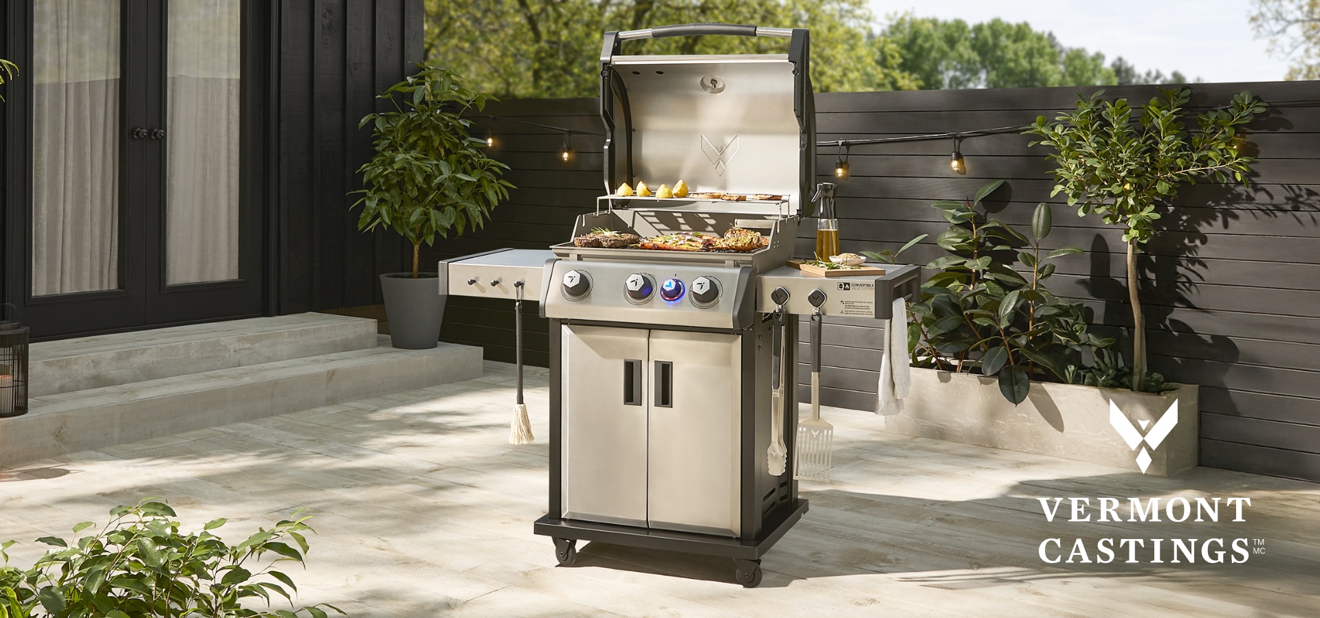 A Vermont Castings BBQ with an open lid, griling various meats and vegetables and dressed with various Vermont Casting accessories on a sunny patio.