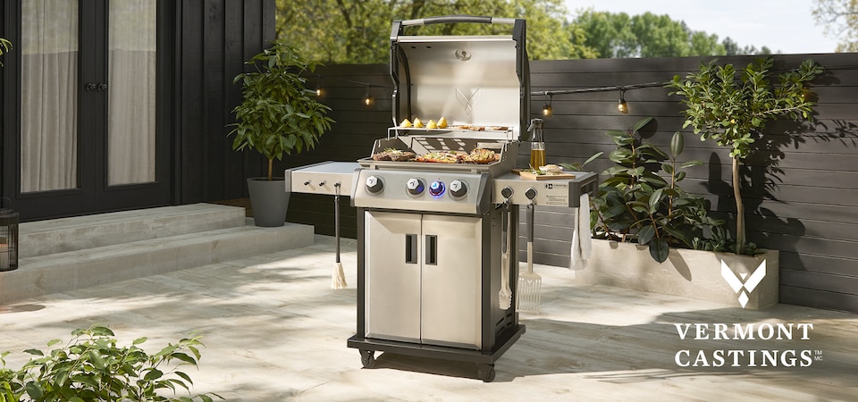 Un barbecue Vermont Castings avec couvercle ouvert, sur lequel grillent divers légumes et viandes et garni de divers accessoires Vermont Casting sur une terrasse ensoleillée.